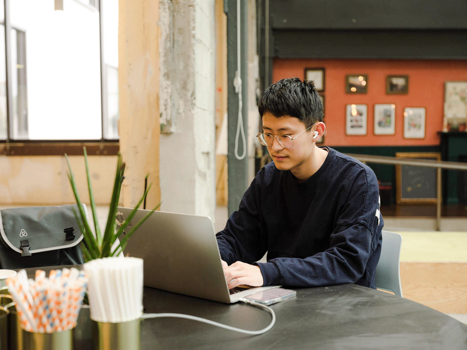 A student wearing earbuds and working on a laptop on a table in the office. There is an Airbnb backpack in the chair next to him.